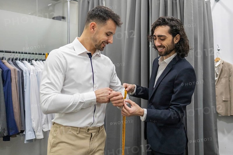 A tailor measures a customer for a custom suit in a suit store. The scene captures precision and professionalism in tailoring, with a range of suits displayed