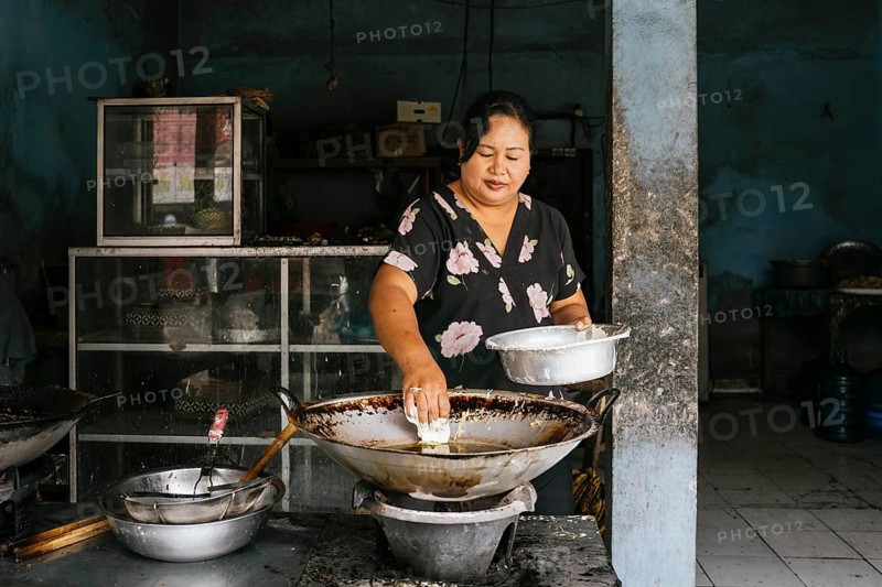 A woman prepares traditional food in a rustic kitchen of Bali, focusing intently as she cooks over a large wok. The indoor setting is simple, with an authentic local atmosphere