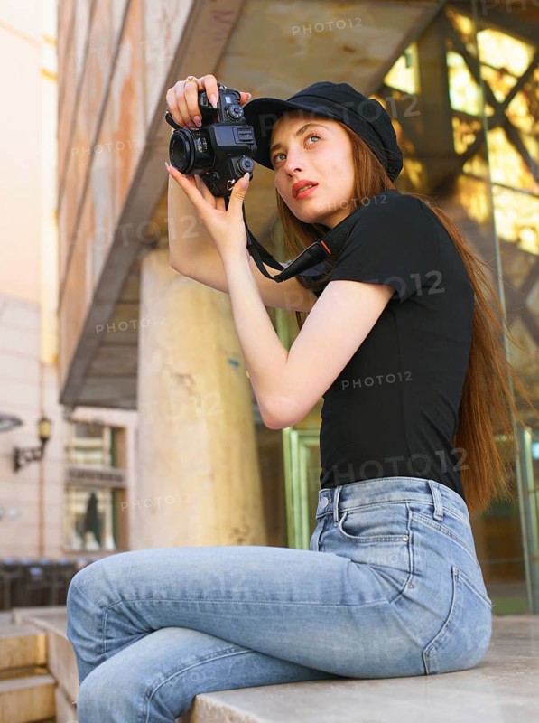 A woman in casual attire takes photos with a professional camera in a city setting. Her focused expression and stylish look reflect her passion for photography and urban exploration