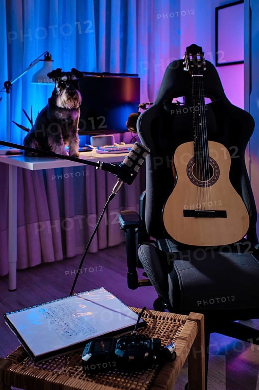 A cozy home studio with a guitar on a chair, a dog on the desk, and ambient purple and blue lighting. A microphone and notebook enhance the creative atmosphere