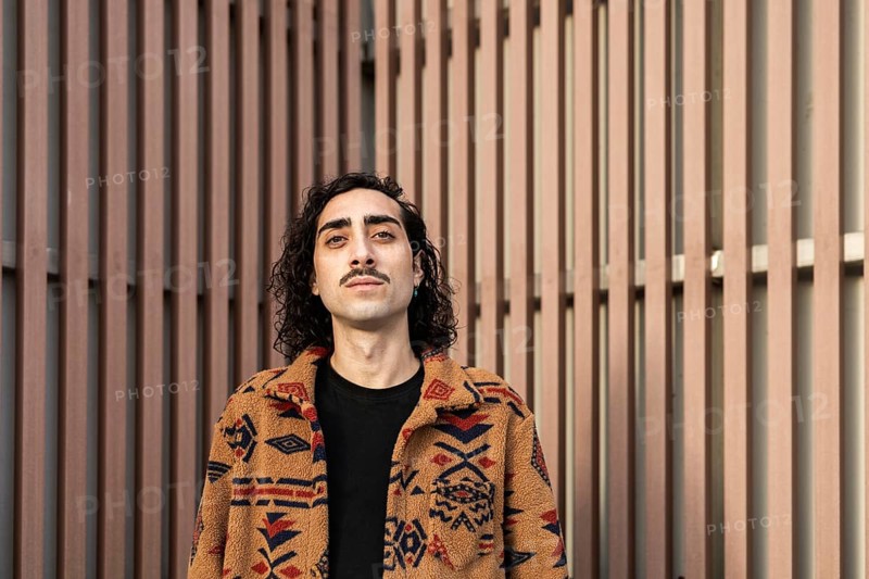 Portrait of a confident man with a mustache, wearing a patterned jacket, standing against a modern beige urban backdrop The image captures a contemporary lifestyle vibe