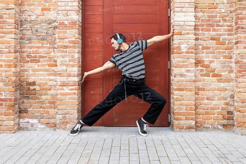 A cuban dancer exudes creativity and style, striking a dynamic pose against a vibrant red door framed by rustic brick. His casual attire and headphones reflect a modern vibe