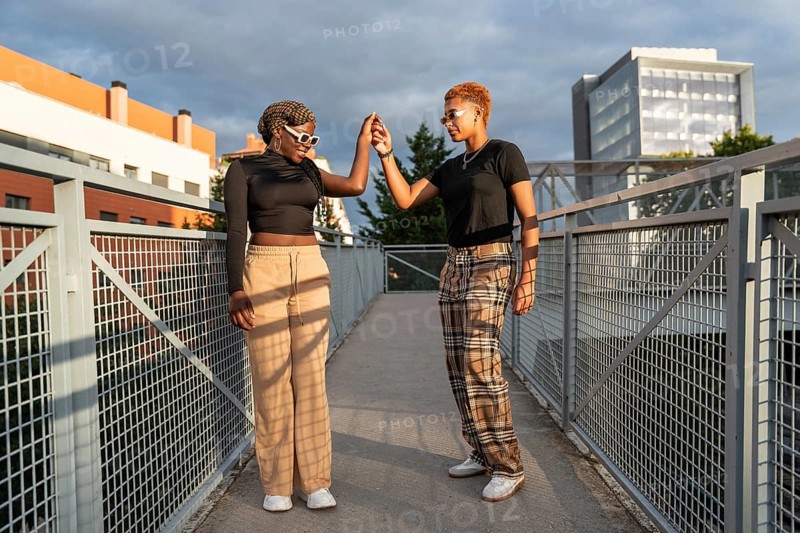 A cheerful LGBT multiethnic couple enjoys a sunny day on an urban bridge, holding hands and smiling, dressed casually. The scene captures warmth and connection against a city backdrop