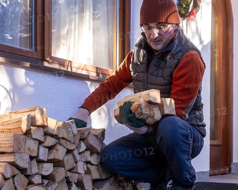 A senior man carefully stacks pieces of eco-friendly firewood outside his home on a sunny day, highlighting sustainable domestic practices and an active lifestyle