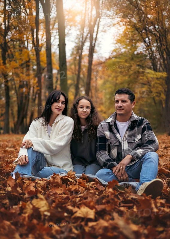 A family sits amidst a forest blanketed in vibrant fall leaves. The setting sun filters through the trees, creating a warm, picturesque autumn scene filled with tranquility