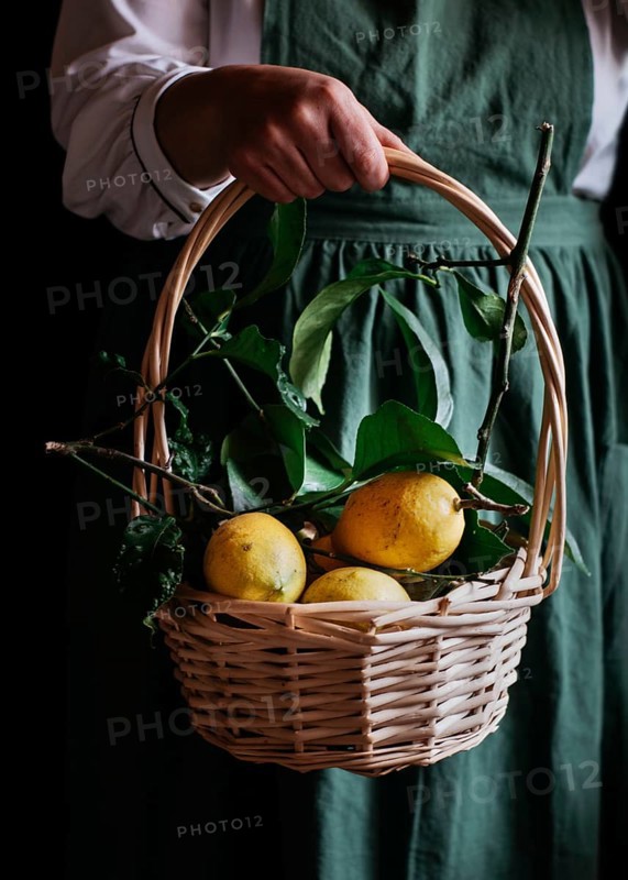 A close up of a wicker basket filled with fresh, ripe lemons, held by a person wearing a green apron. The vibrant yellow contrasts beautifully with the rustic basket