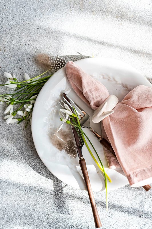 From above, an elegant spring-themed table setting featuring fresh snowdrops arranged on a white plate, complemented by a pink napkin and vintage cutlery, highlighted by soft natural light