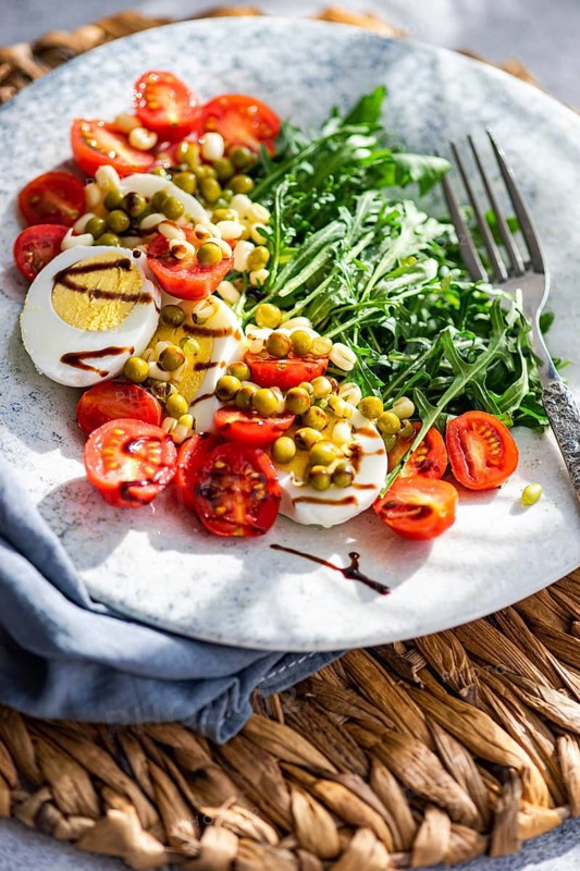 A vibrant keto lunch featuring boiled eggs, fresh rocket leaves, cherry tomatoes, canned green peas, and sprouted green beans, all dressed with balsamic sauce