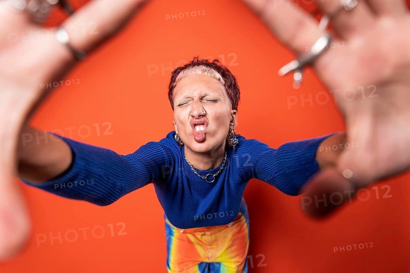 Fashion-forward young woman with vibrant orange background, wearing colorful trousers, tooth gems and a blue sweater. Expressive pose highlights her modern, bold style