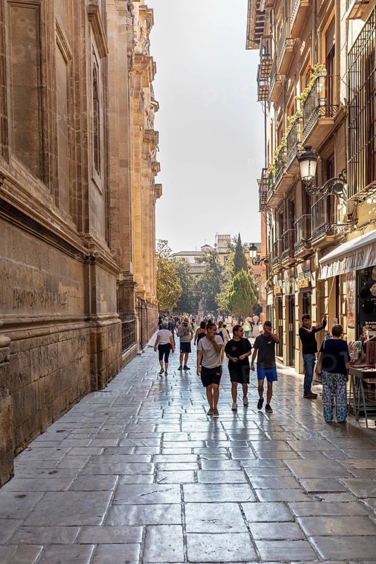 People stroll through a narrow, sunny alley with historic buildings, Granada