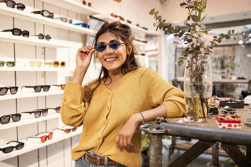 A cheerful Latin woman wearing stylish sunglasses poses in a trendy eyewear shop, showcasing a variety of fashionable frames. Her vibrant smile exudes confidence and style