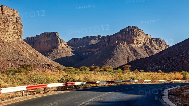 A road with a bend winds its way through impressive mountain scenery in a barren desert landscape under a bright sun, A drive through the mountain landscape of the Sahara in Algeria