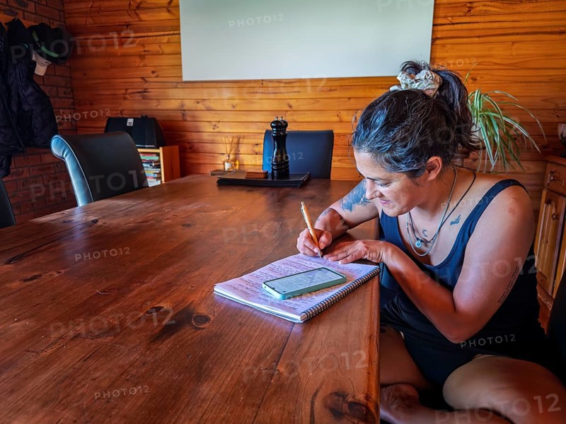 A woman is writing in a notebook at a large wooden table, surrounded by warm decor in Australia. Her phone is nearby, suggesting a blend of digital and analog note taking