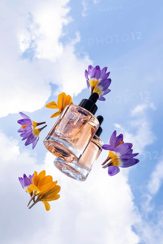 A glass skincare bottle with black dropper cap are showcased among colorful flowers, set against a bright, cloudy sky. A perfect blend of elegance and nature beauty