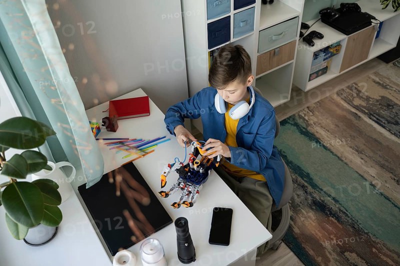 A young boy sits at a white desk, focused on assembling a robot. Surrounded by colorful tools, headphones around his neck, he's absorbed in a creative and educational activity