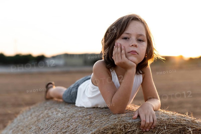 Child in casual attire lies on a hay bale during sunset, reflecting a calm and serene mood. The warm sunlight enhances the peaceful rural atmosphere