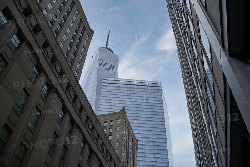 An upward angle captures the iconic silhouette of the One World Trade Center amid surrounding buildings in Manhattan, against a blue sky