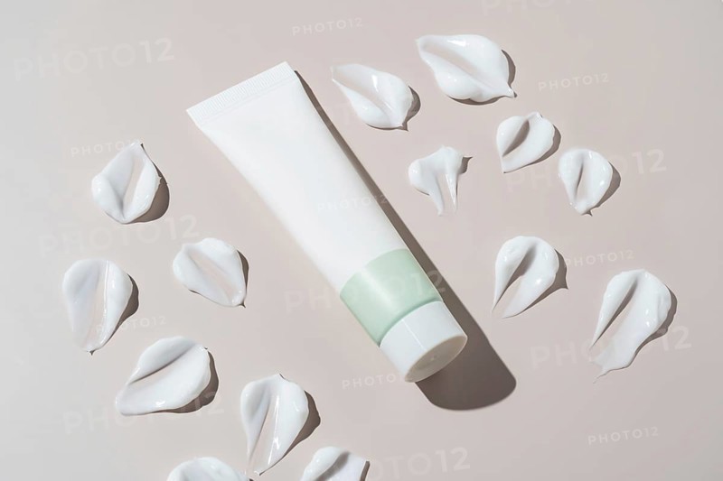 An elegant cosmetics container with a minimalist design appears next to artistic cream stains on a neutral background. The textures add depth and elegance to the composition