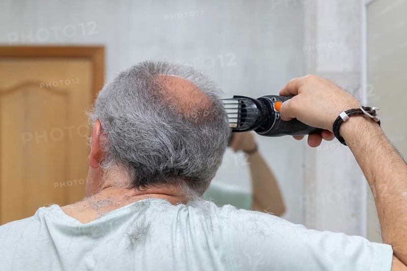 A man cuts his gray hair with electric clippers. The scene captures a home-style haircut in a casual, homey setting