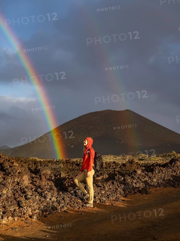 A woman in a red jacket stands on volcanic terrain in Timanfaya National Park, Lanzarote. A vivid rainbow arches across the sky, highlighting the contrasting natural beauty