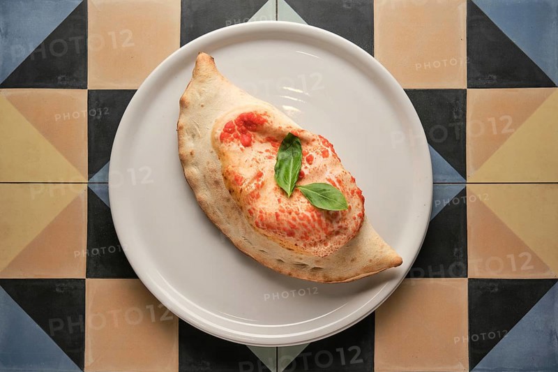 Freshly baked calzone garnished with basil leaves, served on a white plate over a vibrant geometric tiled surface