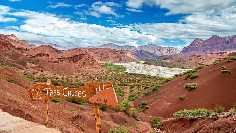 Red mountain landscape with vegetation and a sign in the foreground under a blue sky, The landscape and colorful rock formations of the Quebrada de Cafayate near Salta in Argentina