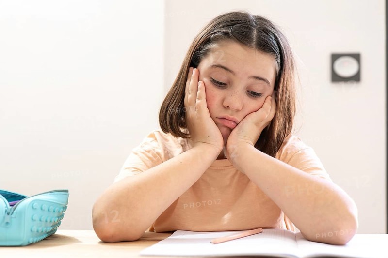 A young girl sits at a desk, appearing stressed and overwhelmed with her homework. Her head rests on her hands, and a pencil case sits close by, evoking school life challenges