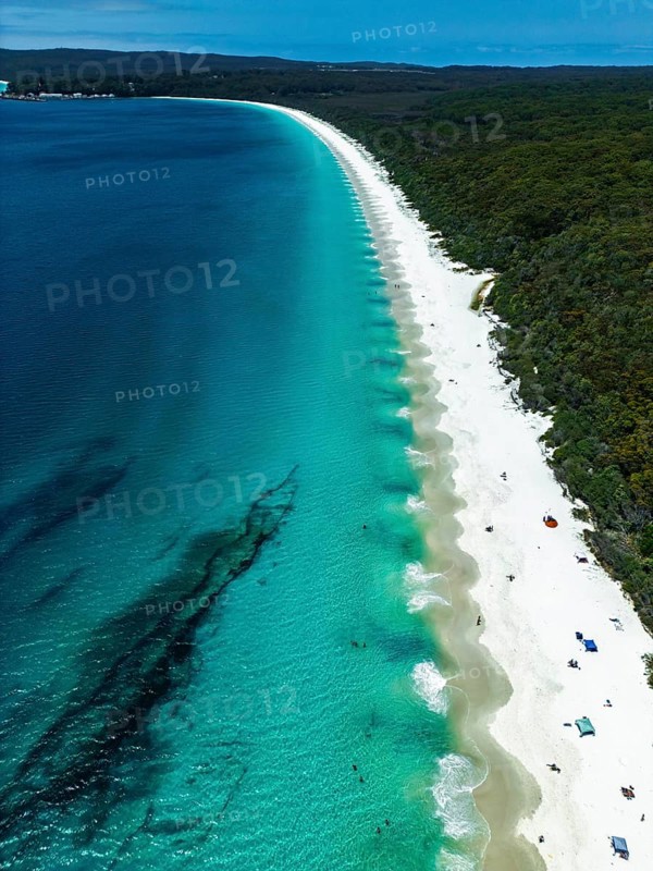 Aerial view of a pristine beach with vibrant turquoise waters and white sand in Jervis Bay, Australia. Dense greenery lines the shore. Perfect getaway for sunbathing, swimming, and nature