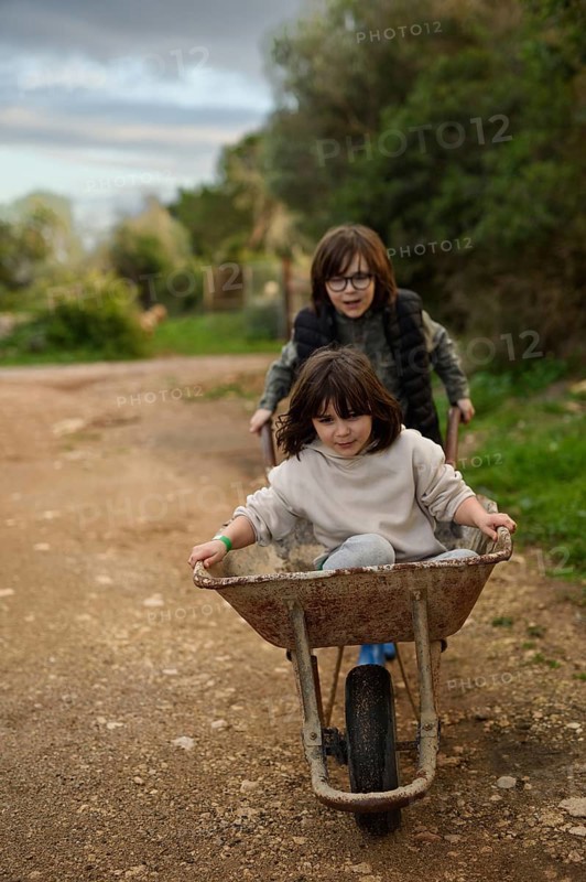 Two children enjoying a playful moment with a wheelbarrow on a rural path. The older child pushes while the younger one rides, surrounded by lush greenery and open space
