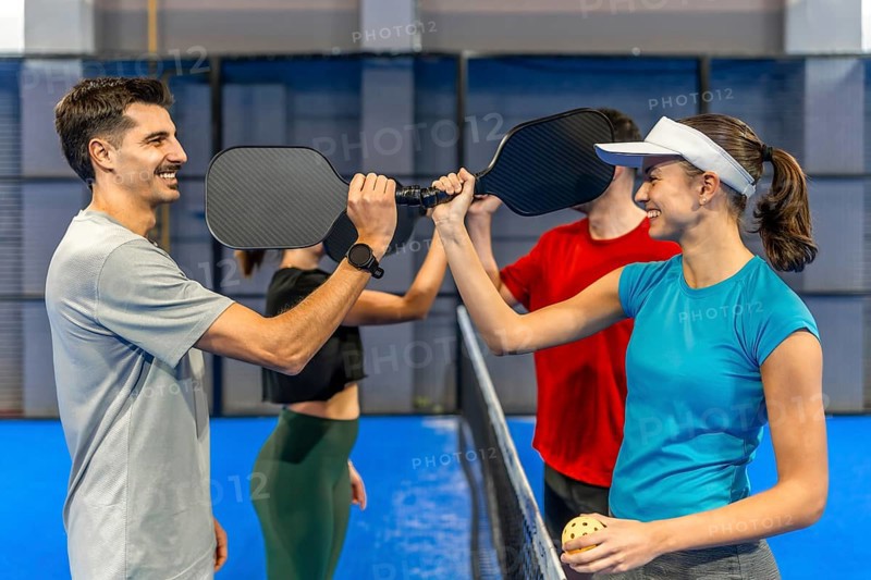 Four friends on a blue pickleball court paddle to paddle over the net, smiling and high fiving after a friendly match, celebrating teamwork, fitness, and fun