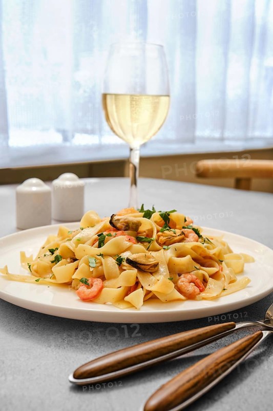 A plate of seafood pasta with shrimp and clams on a kitchen table. A glass of white wine stands nearby, complementing the meal in a bright and inviting dining space during midday
