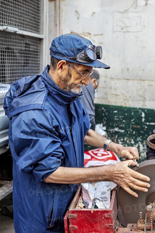 A man working on a machine in a workshop, wearing a blue jacket and a cap, A man sharpening knives in the historic centre of Algiers Algeria