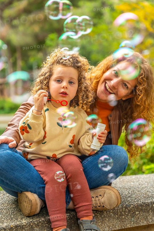 Vertical portrait of a cute blonde girl and mother sitting on a park blowing bubbles