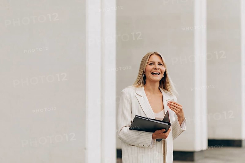 A businesswoman in a white suit smiles joyfully while holding a folder. She stands in front of sleek architecture, capturing a professional and optimistic demeanor
