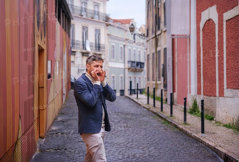 Man musician in a blue blazer and white sweater playing blues on a harmonica with eyes closed, standing outdoors in city street