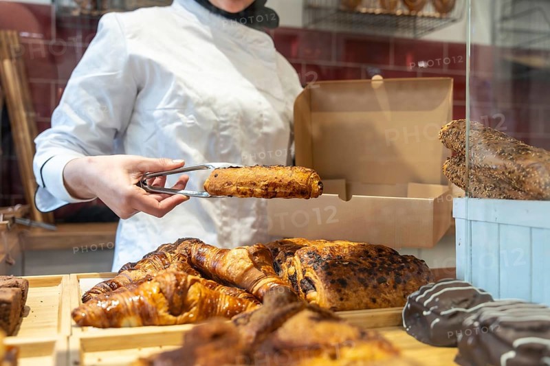 Bakery worker in uniform uses tongs to place a warm pain au chocolat into a paper takeaway box amid a tempting display of fresh pastries and bread on the counter
