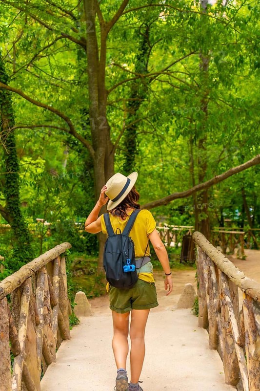 A woman wearing a straw hat and a green backpack is walking across a bridge. The bridge is surrounded by trees, and the woman is enjoying her walk