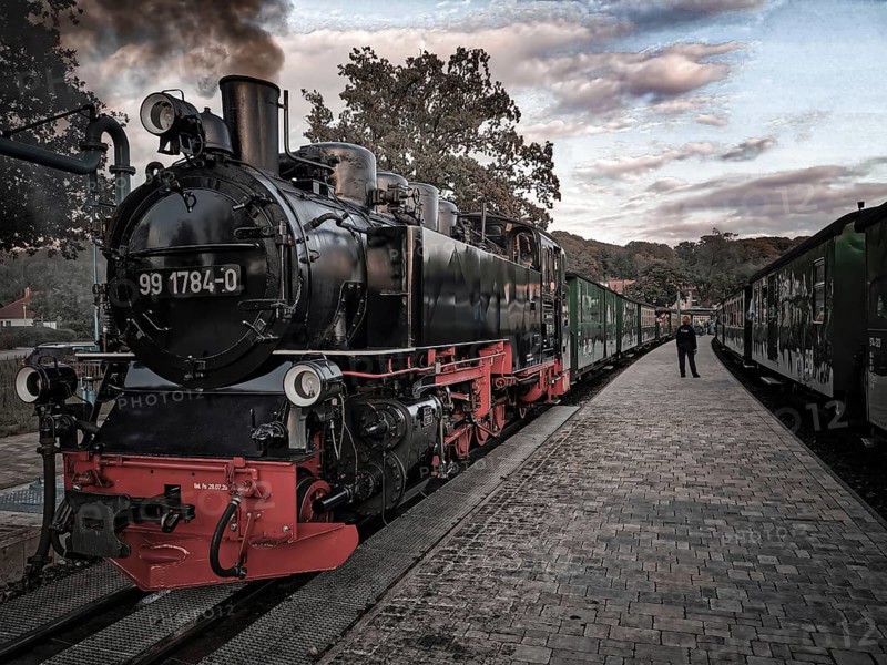 Old, historic steam locomotive in bright colors on tracks at the station in a nostalgic atmosphere, front view of speeding Roland on Rügen at sunset
