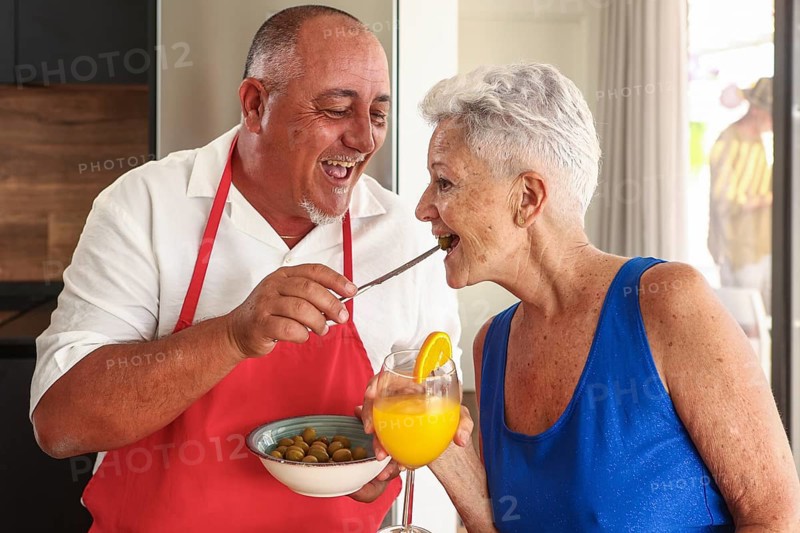 A joyful senior couple engages in a playful and affectionate interaction in a bright kitchen during a party, sharing olives and orange juice