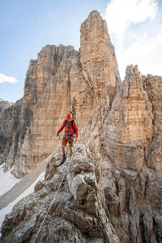 Mountaineer climbs on an exposed rock in the secured Via Ferrata Bocciere Centrale via ferrata, mountain landscape with spectacular rock towers in the back, Brenta-Adamello Nature Park, Trentino, Italy