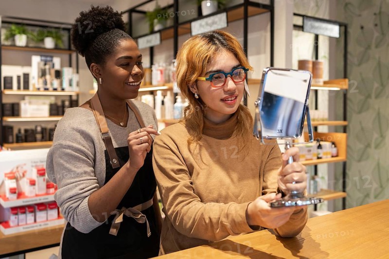 A friendly cosmetics store worker assists a customer trying on products. The customer holds a mirror, reflecting a vibrant store ambiance focused on personalized beauty service
