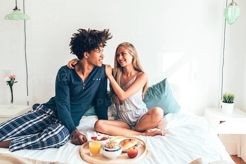 A young couple sits cheerfully on a bed, sharing a breakfast tray filled with fruit, juice, and pastries. The room is well-lit, creating a warm, intimate atmosphere