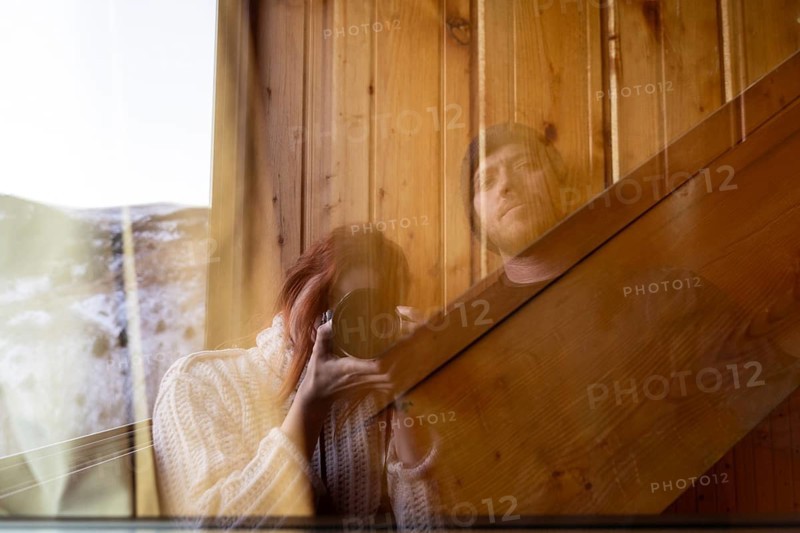 A woman with red hair holding a camera and a man are reflected in the glass window of a rustic cabin, symbolizing a serene and intimate winter moment