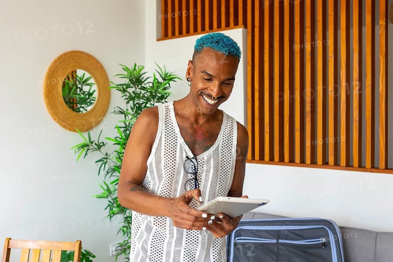 Hispanic LGBTQIA+ man with blue hair happily prepares for holidays using a tablet, surrounded by cozy home decor and greenery. Embracing celebration and diversity
