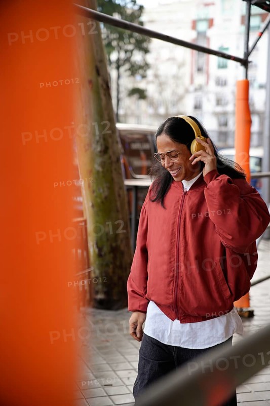 A man with long hair and glasses enjoys music through yellow headphones while walking outdoors in a red jacket, exuding happiness in an urban setting