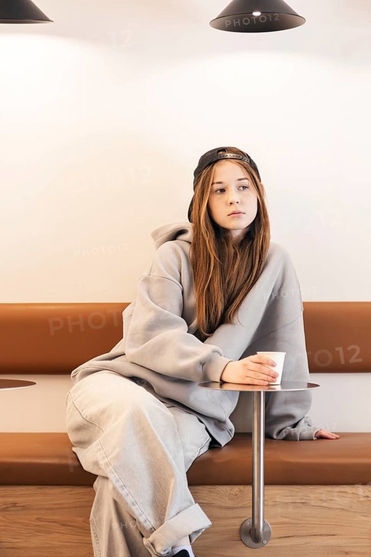 Teenage girl in casual attire with a cap, sitting thoughtfully on a bench. She holds a paper cup, relaxed and contemplative in a minimalist indoor setting