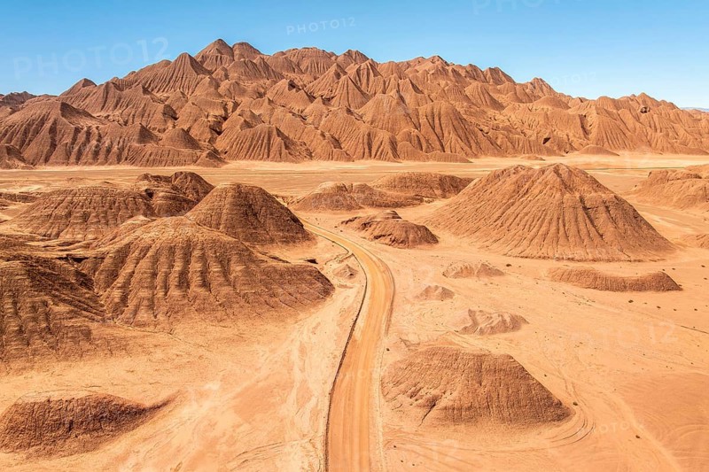 Aerial view of a vast desert landscape featuring a rugged mountain range and a winding dusty road under a clear blue sky in the argentine Puna, showcasing the beauty of arid terrains