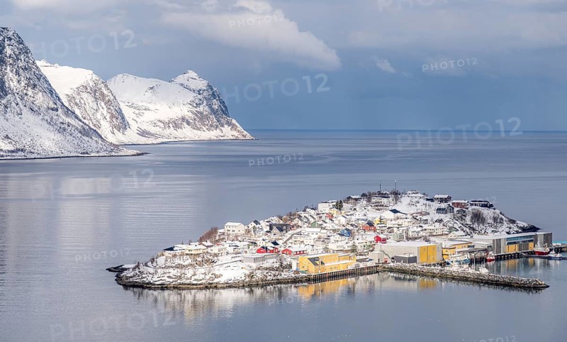 Scenic winter view of Husoy, Norway, with snow-covered houses nestled between serene waters and majestic mountains, creating a tranquil and picturesque coastal landscape
