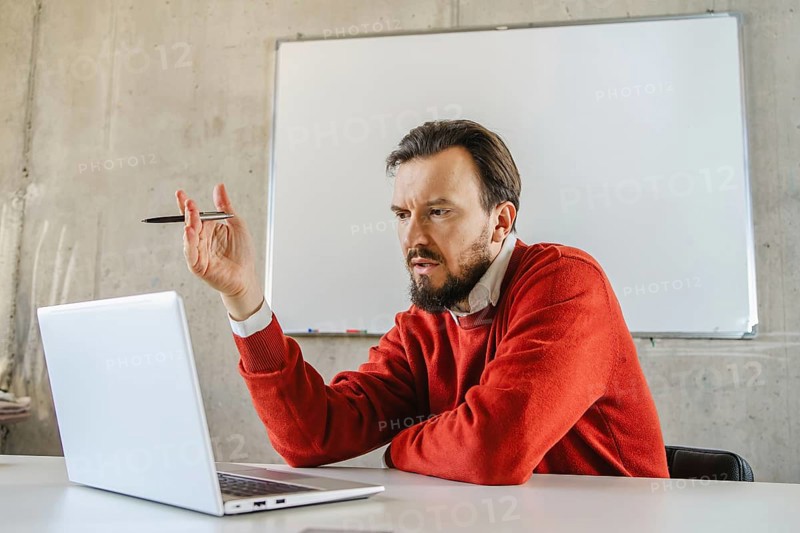 A office man in a red sweater gestures expressively during a virtual meeting. He is seated at a desk with a laptop and whiteboard, embodying remote work dynamics