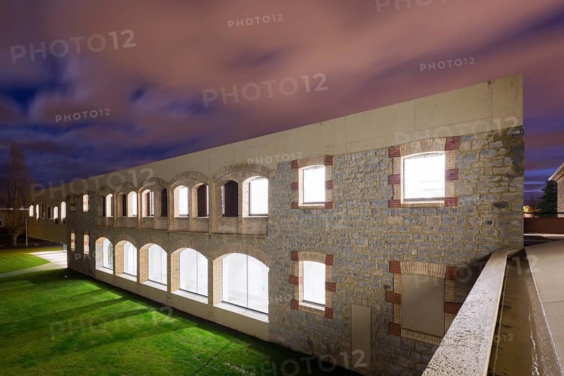 A beautifully illuminated stone hospital with arches under a twilight sky, showcasing architectural elegance. Green grass contrasts with the vivid sky, capturing serenity